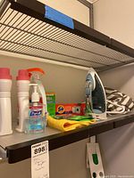 Shelf showing assorted cleaning supplies including Purell hand sanitizer, spray cleaner, cleaning cloths, Tide and Affresh cleaning products, a Black & Decker iron, and a laundry bag.