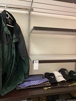 Eddie Bauer men's jacket hanging on a white hanger, predominantly green with darker upper part and fleece collar. Below jacket is a black travel case containing several ties, and on the shelf below are four caps and some gloves alongside a shoehorn.