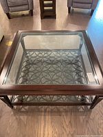 Square coffee table seen from above showing clear glass top, dark wood frame, and decorative metal pattern under glass.