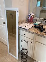 Photo of the full-length white framed wall mirror and metal towel tray on floor beside bathroom cabinet.