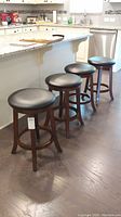 Four dark wood framed bar stools with round black leather seats placed in a kitchen at counter height. Floor is dark wood. Counter has white and grey granite top.