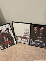 Set of three posters with titles Django Unchained, Forrest Gump, Goodfellas, framed in black plastic frames and resting against a wall on carpeted floor.