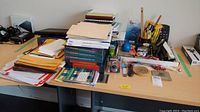 Wide view of table with stacks of papers, folders, notebooks, office stationary, and tools all assorted and piled.