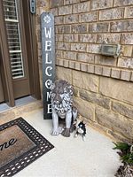 Resin lion statue, small dog statue, and welcome sign positioned near a brick wall and a door mat outdoors.