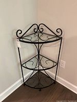 Full view of the black iron corner shelving unit with three glass shelves in a corner of a room on hardwood flooring, showing decorative curled ironwork on top.