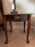 Front view of a round wooden side table showing curved legs and a drawer with brass hardware.