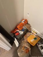 Metal Sears Craftsman toolbox with orange extension cord coiled on top and miscellaneous bags containing repair materials and tools around it, placed in corner on floor.