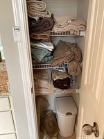 Closet shelf with stacks of folded towels in brown and beige, folded bed linens, and white plastic trash can on floor.