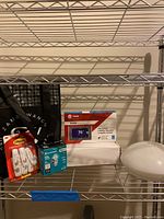 Wide view of the metal shelving with all items including Trane thermostat box, ceiling light globe, two packages of light bulbs, and Command hooks.