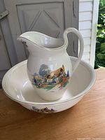Full view of antique ceramic pitcher and bowl set on wooden surface with outdoor background. Pitcher sits inside bowl. Both pieces white with painted cottage scene.