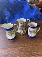 Photo of three antique English pitchers on wooden table showing floral, farm scene, and decorated handles