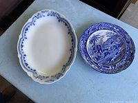 Both ceramic pieces displayed on a surface showing size and shape difference, platter larger and oval, plate smaller and round with patterned decoration