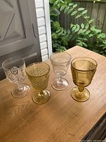 Four antique Nova Scotia goblets on wooden table, showing two amber and two clear glass goblets with ornate patterns and stems, photographed on outdoor table.