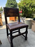 Single oak chair with worn brown leather upholstery and brass studs around seat and backrest edges, showing wooden 'sausage' turned legs and stretchers.