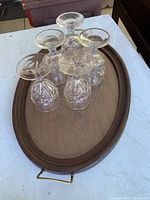 Top-down view of 5 glass water goblets sitting upside down on the oval wooden serving tray with brass handles