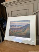 Framed pastel landscape artwork resting on a wooden surface, showing layered mountains and sky in pastel colors, signed by the artist.