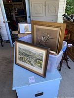 Photo showing two framed artworks at forefront including vintage landscape print and floral painting with additional frames in plastic bin behind