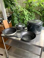 Photo showing six kitchen cookware items arranged on a stainless steel table: wok pan, covered pot, pot with sieve, and large canning pot