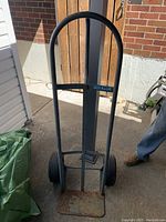 Full front view of a metal hand truck dolly showing black rounded handle, vertical frame, worn rusty base plate, and two wheels on floor.