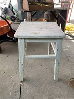 Front view of antique pine bench with square top and visibly splayed legs resting on a concrete floor.
