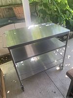 Full view of the steel dolly cart showing the three metal shelves, handle, and wheels, placed outdoors on a concrete floor.