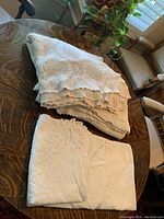 Three vintage cotton bed covers folded on an antique wooden table, showing delicate crochet patterns and embroidery details.