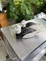 Two vintage duck decoys placed side by side on a metal surface with greenery in the background, showing overall size, shape, and paint wear.