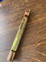 Polished red brass thermometer laying on a wooden surface, showing full length with hanging loop and temperature scale.