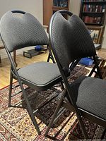 Photo showing three folding chairs with gray padded seats and backs, metal frames, placed on a patterned rug in a living room.