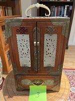 Front view of antique wooden jewelry box showing two doors with jade carved panels, brass decorative hardware and handle on top.