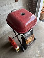 Closed red kettle BBQ grill standing on a black wheeled stand with a bag of charcoal and two condiment bottles placed beside it.