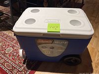 Full exterior front view of the Coleman cooler showing blue body, white lid with four cup holders, and attached wheels.