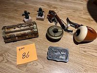 Photo of 9 collectible brass, lead, and wood craft items arranged on wooden floor including pipes, figurines, compass, carved box, and plaque.