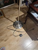 Floor lamp base showing weighted round metal base and attached electrical cord with foot pedal switch lying on hardwood floor.