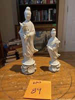 Photo showing two white porcelain Guan Yin sculptures on a wooden surface, with one taller and one shorter figure both holding lotus flowers.