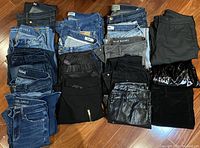 Sixteen pairs of jeans and skirts folded and stacked on floor under direct light showing various colors and brands