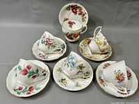 Six sets of white porcelain teacups and saucers each with colorful floral patterns and matching saucers, arranged on a flat surface.