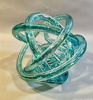 Front and side view of a modern aqua blue glass knot sculpture on a white surface.
