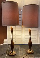 Pair of mid-century modern teak and brass table lamps with light brown fabric shades showing wear.