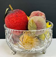 Front and side view of crystal footed fruit bowl filled with red, purple, yellow, and peach colored fake fruits showing textured surfaces and fabric leaves.