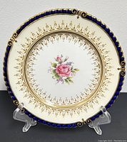 Single 6.25″ dessert plate displayed upright on stand, showing cobalt blue scalloped edge, gilt bead border, pierced lattice and central pink rose