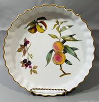 Full top view of the pie plate showing scalloped edge with gold trim and colorful fruit illustrations of peaches, blackberries, and apple halves.