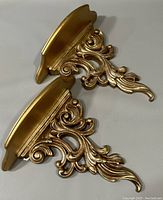 Top angle view of the two gold ornate vintage wall shelves laid flat, showing the detailed scrollwork design and curved shelf tops.