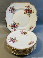 Close-up photo of stacked square dessert plates showing floral clusters with pink, yellow, and blue flowers and gold rims.