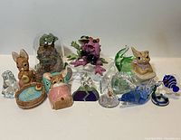 Full view of thirteen assorted figurines including Pendelfin rabbit ceramic figurines and various colored glass animal and decorative figurines arranged on flat surface.