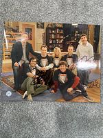 Photo shows six cast members of The Big Bang Theory in a living room setting with multiple autographs on the photo surface.