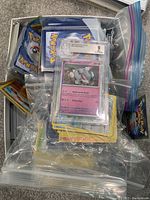 Top-down image of a box filled with plastic bags containing numerous Pokémon cards, showing a prominently graded Greenard card.