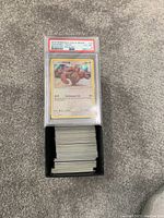 Photo showing a PSA graded 2019 Pokémon Sun & Moon Lickitung holo card graded EX-MT 6 on top of a black container holding a large bulk of loose Pokémon cards.