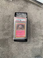 Image showing a stack of Pokémon cards on carpet floor with the top card being a 2018 Sun & Moon Jigglypuff holographic card graded PSA GEM MT 10 inside a plastic protective case, with additional bulk cards beneath.
