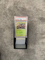 PSA graded Lurantis card on top of a large stack of ungraded Pokémon cards in a black box on carpet.